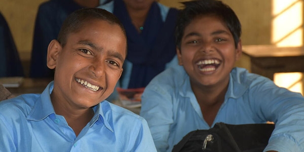 Two boys wearing school uniforms sit together at a desk at a school in Nepal and laugh.