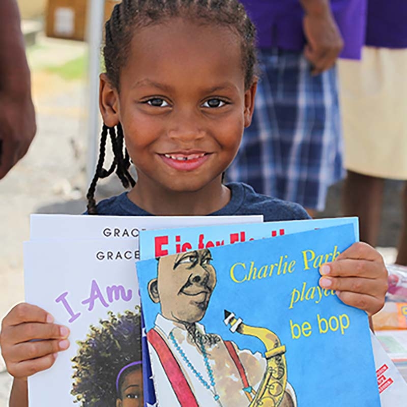 A young girl holds three pictures books and smiles. 