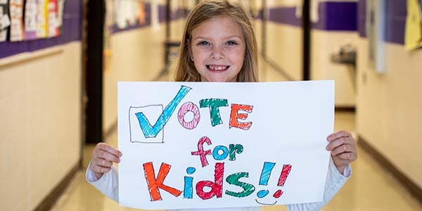 An elementary student smiles while holding a sign that reads, Vote for Kids.