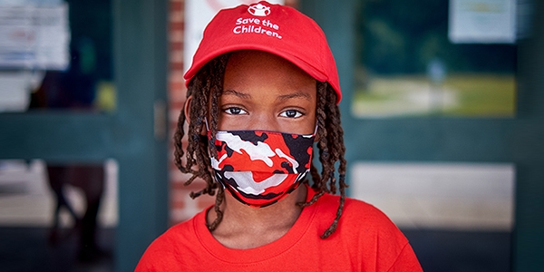 A boy wears a red Save the Children hat and a cloth face mask while standing outside.