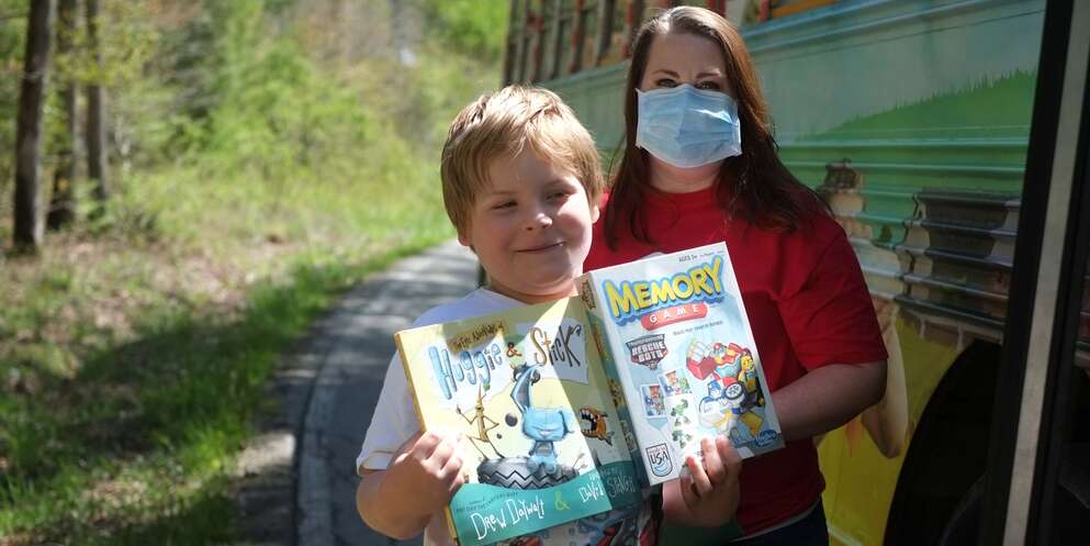 A Save the Children staff member stands near a school bus wearing a face mask as a young boy holds two picture books next to her.