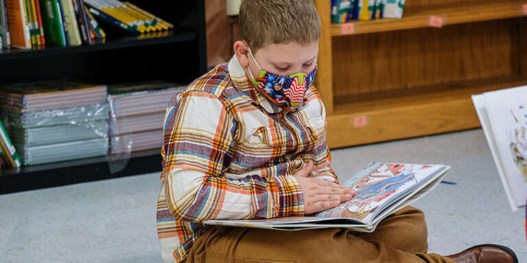 A boy sits on the floor reading a book and wearing a mask. 