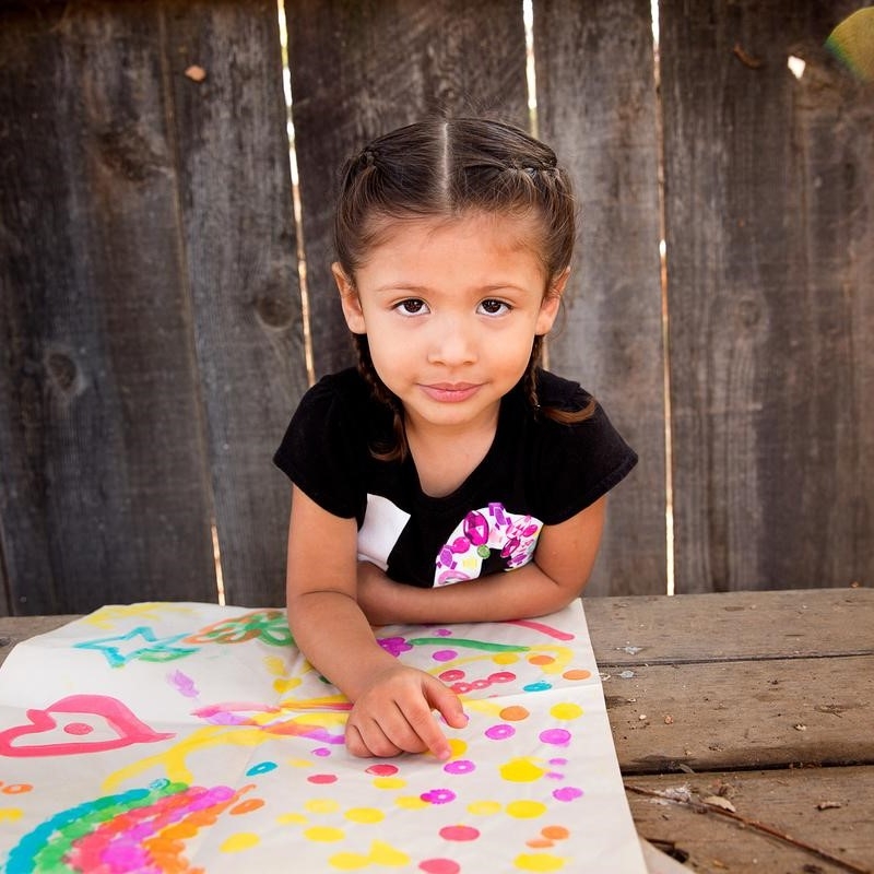 A 3-year old girl named Analia sits at a picnic table outside and displays a colorful painting in front of her. The girl and her mom participated in several learning-based activities taught to them during a home visit as part of Save the Children’s signature Early Steps to School Success program. Photo credit: Save the Children, Nov 2017.