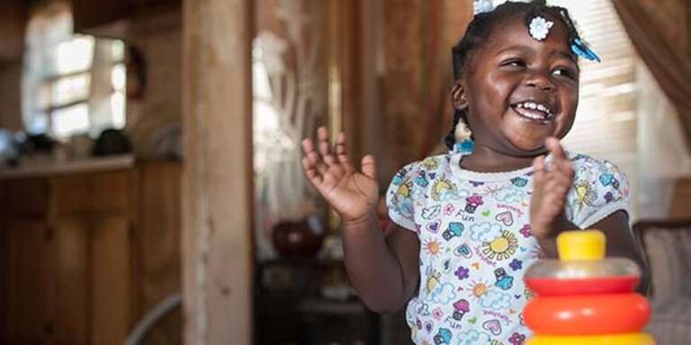 A 20-month old girl who lives in Mississippi smiles as she gets ready to clap her hands while standing near a table with a bright toy on it.