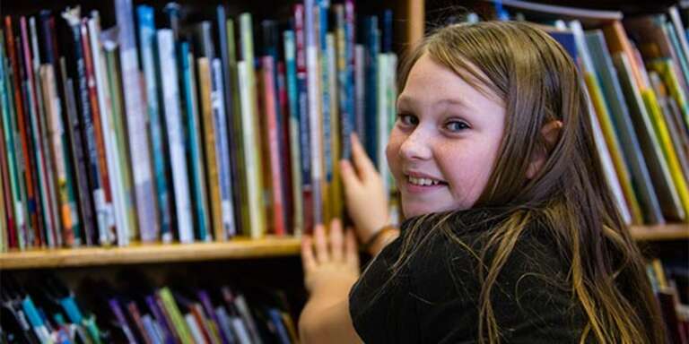 A young girl in a library smiles at the camera while taking a book off the shelf. 