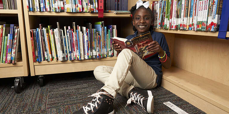 A girl named Riley sits on the floor of her school’s library and smiles and she holds a thick hardcover book open. The casual reading spot contains dozens of other colorful books, encouraging 3rd grade readers like Riley to read to learn. Photo credit: Michael Nylif/ Save the Children, May 2019.