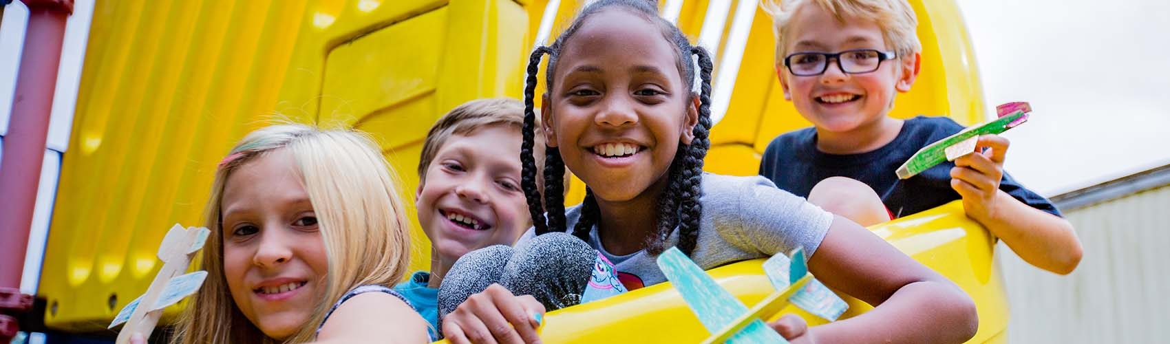 A group of children smile as they gather together on a yellow playground slide. 