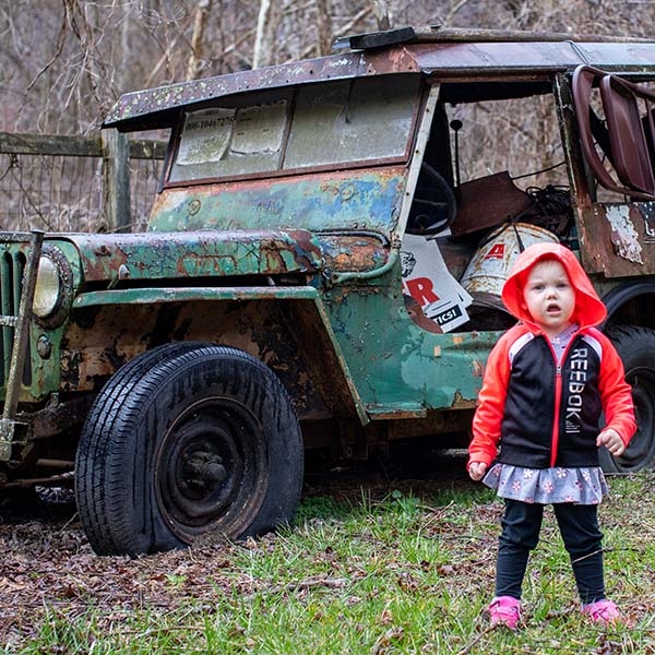 In West Virginia, a girl stands outside in the grass near a rusted tracker. 