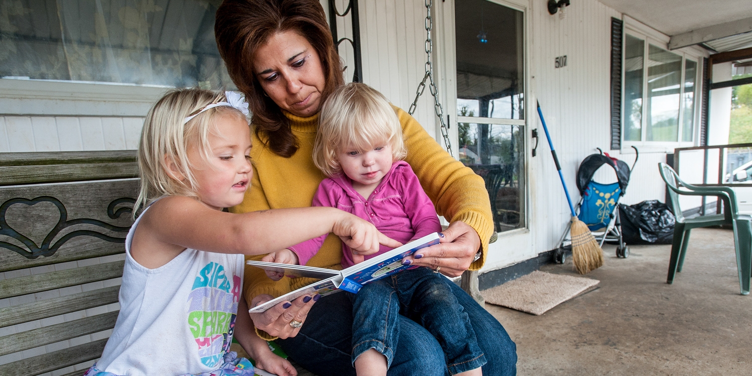 A woman and two children read a book together on their porch.