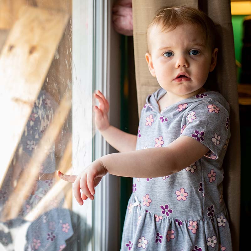A toddler girl holds her palm against a glass window while standing inside her family's West Virginia home.