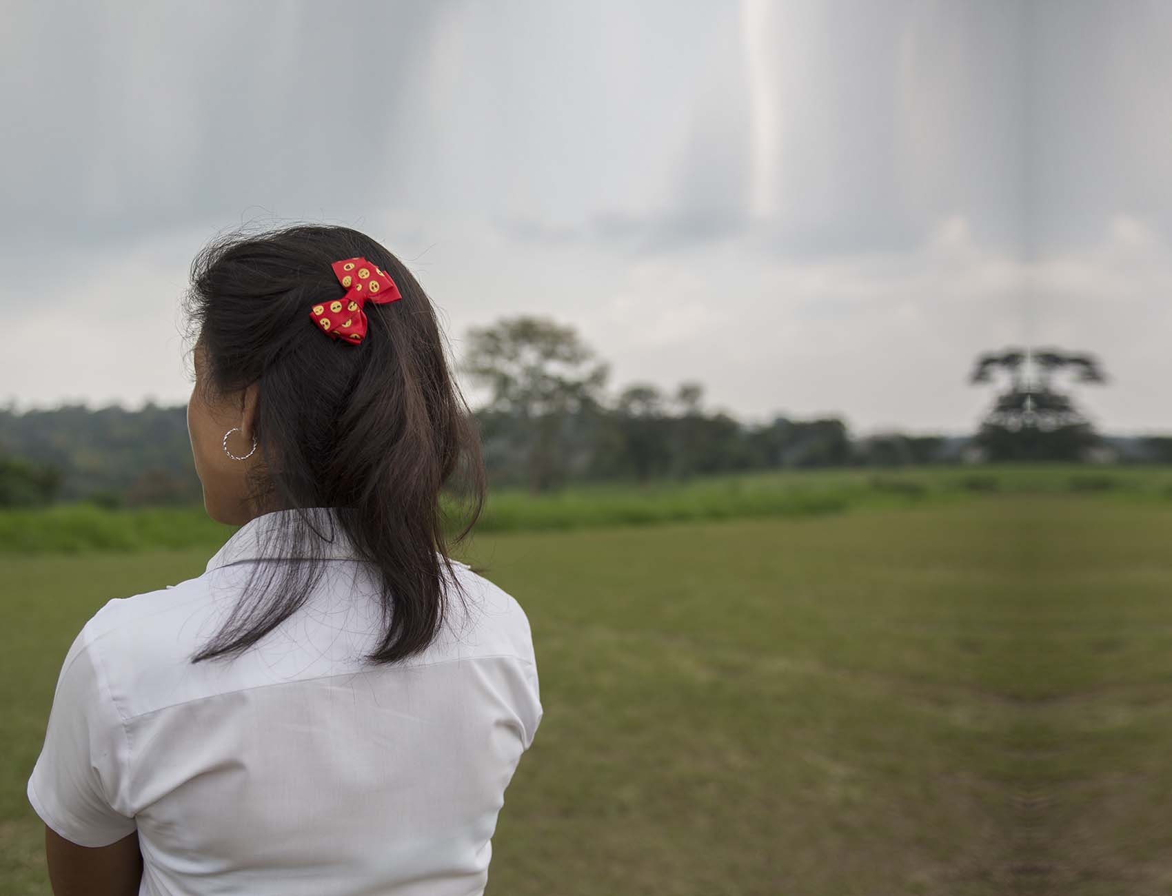 A 14-year old girl stands with her arms cross while looking out over a grassy field in El Salvador. 