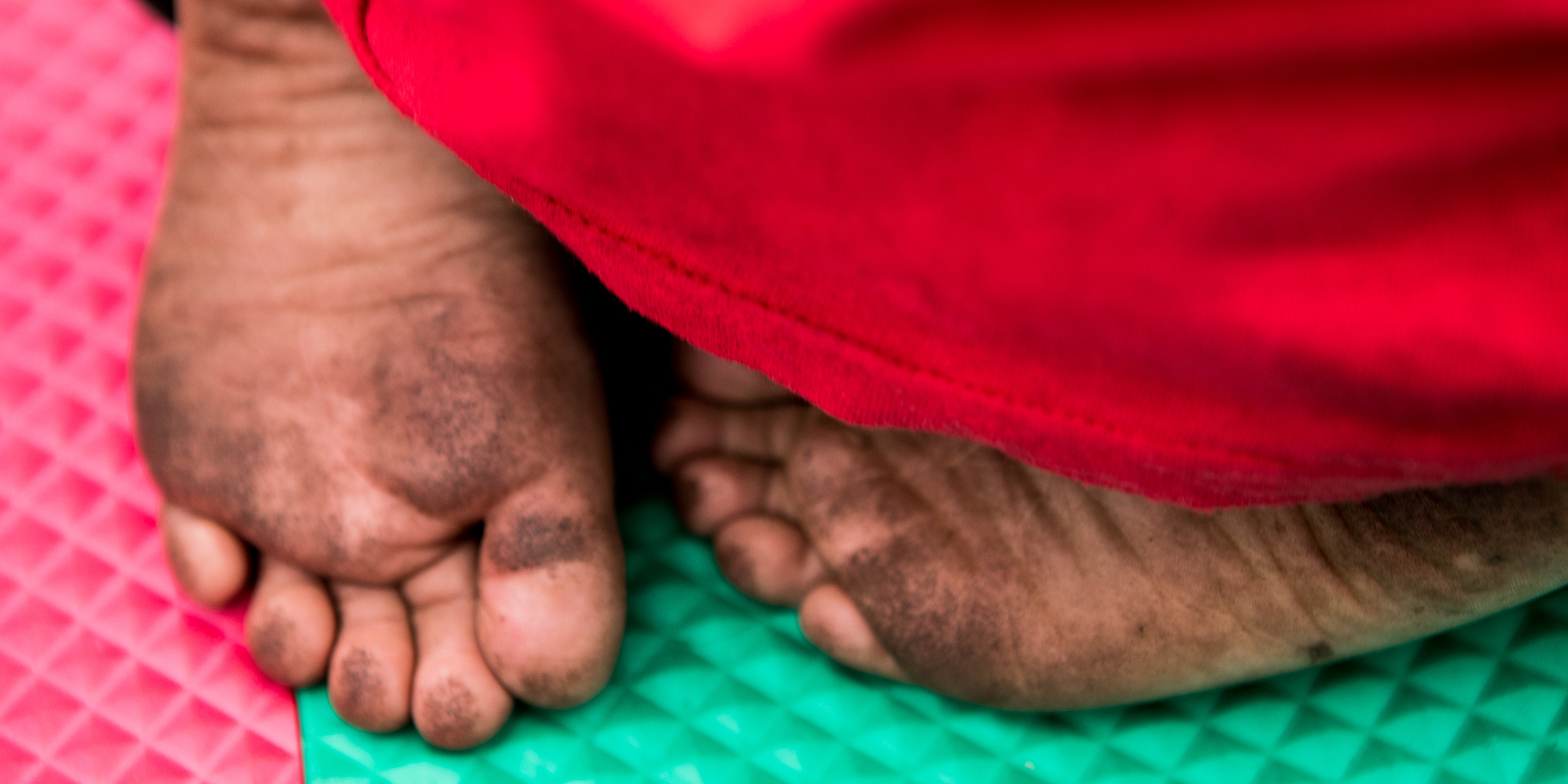The tiny feet of a barefoot child resting on a colorful play mat in a child-friendly space are visible under a red blanket. The child’s feet are covered in dirt. Some children and families crossing the U.S. border have walked for several days and have not bathed, are hungry, sick and exhausted. Save the Children is running child-friendly spaces and children’s play areas at transit shelters in New Mexico for children once they leave detention facilities. Photo credit: Caroline Trutmann Marconi/ Save the Children, Nov 2018. 