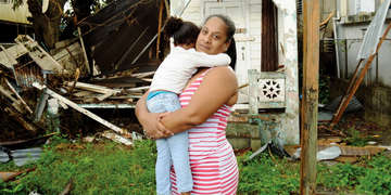 A mother holds her daughter in her arms while standing in front of destruction caused by  Hurricane Maria in Puerto Rico during the 2017 hurricane.