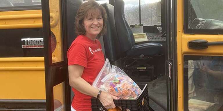 A Save the Children worker holds a crate of food and stands in the doorway of a schoolbus.