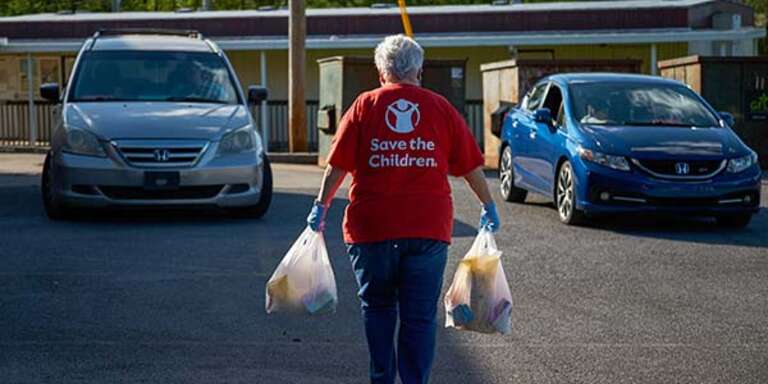 A woman in a Save the Children shirt carries a shopping bag of supplies in each arm. 