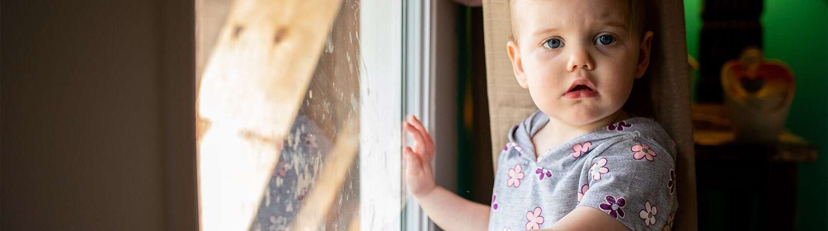 A toddler stands with her hand on a glass sliding door inside her home in West Virginia.