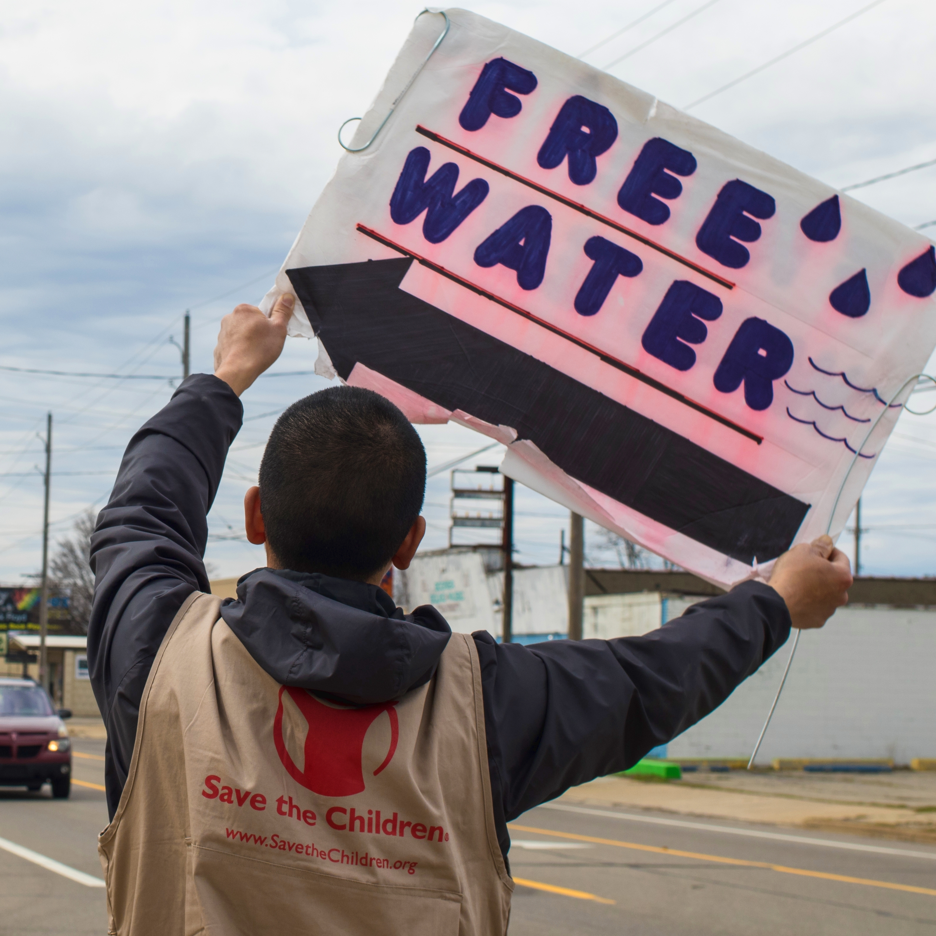 Hee Tae holds a “Free Water” sign up at the intersection to direct people to the water distribution point at NOW Ministry Church in Flint, MI. Save the Children staff in Flint, Michigan assisted the NOW Ministry Church with a daily water distribution to members of the community. Photo Credit: Save the Children/Stuart J. Sia 2016.