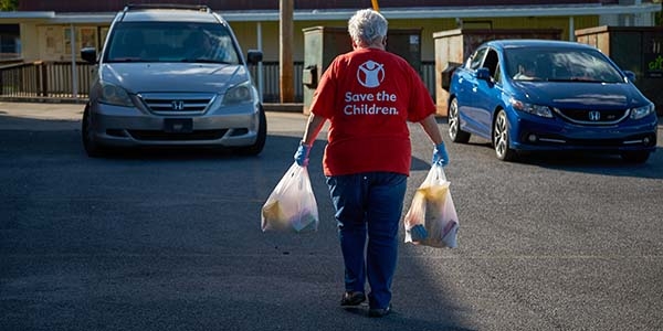 A woman walking away from the camera wears a Save the Children shirt and carries two plastic grocery bags.