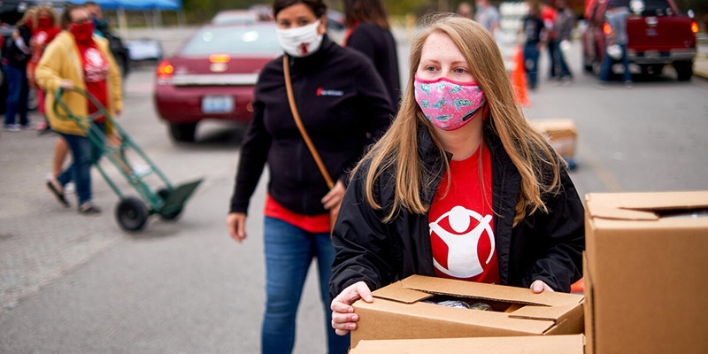 A Save the Children staff member in a red shirt holds a cardboard box of food to distribute to a rural community. 