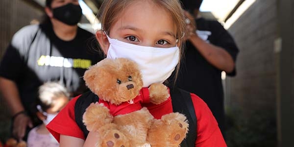 A young girl wears a white face mask while holding a toy teddy bear at a Save the Children distribution center. 