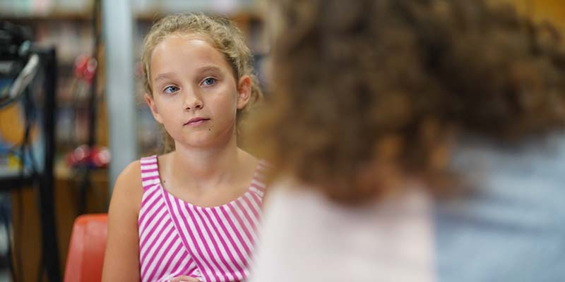 A young girl listens during a conversation while sitting at a table in a school library.