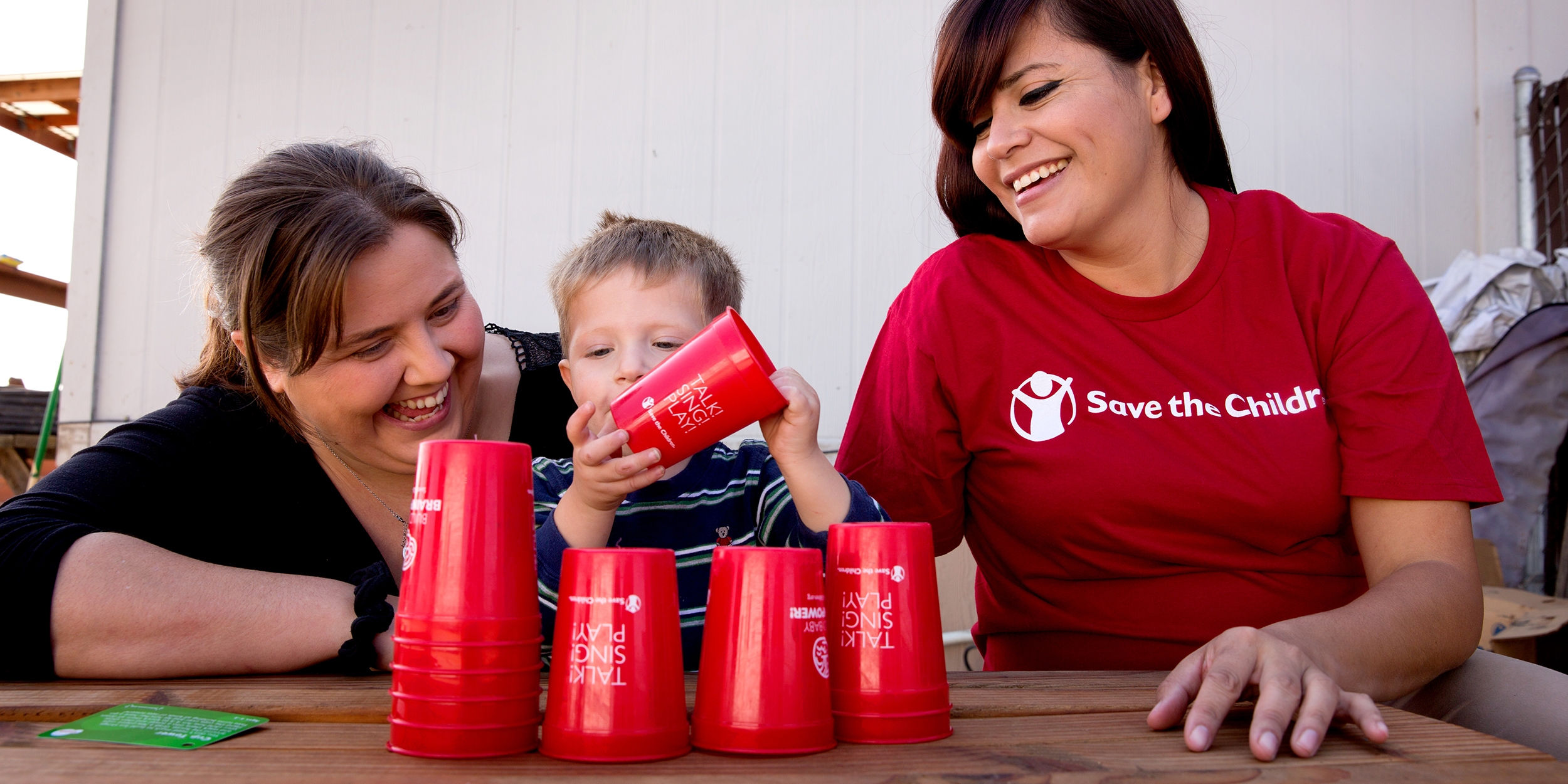 A Save the Children staffer sits at a table while a mother and her child enjoy a game with red cups. 