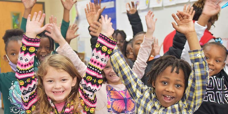 Children smile for a photo with their arms up, excited to play and sing a song.