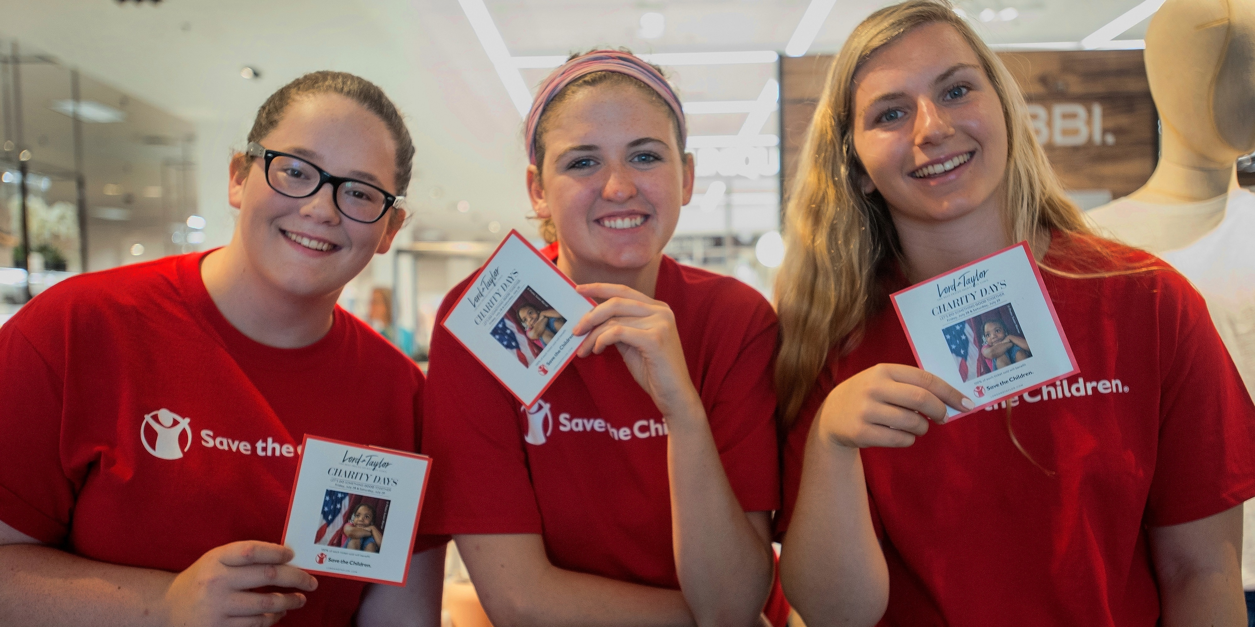 Three women wearing red Save the Children shirts and holding campaign cards gather together at a Save the Children store event. The women are volunteers helping to promote a campaign throughout the store. Photo credit: Susan Warner/Save the Children, July 2017.