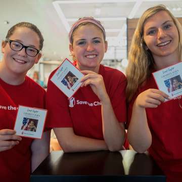 Three women wearing red Save the Children shirts and holding campaign cards gather together at a Save the Children store event. The women are volunteers helping to promote a campaign throughout the store. Photo credit: Susan Warner/Save the Children, July 2017.