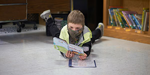 A boy in a camouflage neck gaiter lies on his stomach on the floor of a classroom and is reading a book. 