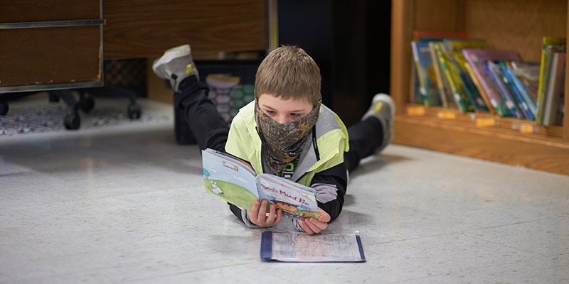 A boy in a camouflage neck gaiter lies on his stomach on the floor of a classroom and is reading a book. 